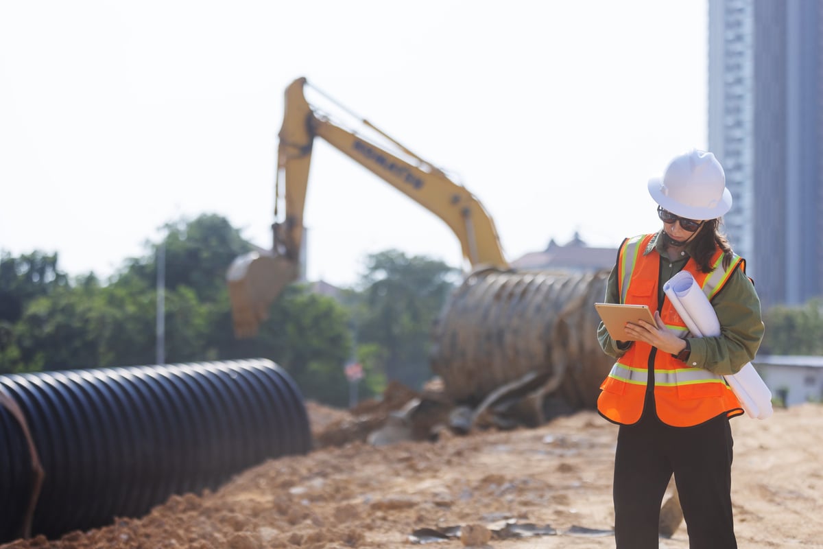A site engineer in a hard hat and safety vest reviewing blueprints and a tablet on a construction site
