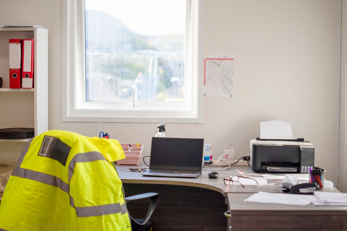 A desk with a laptop, printed documents and a high-visibility jacket hanging nearby